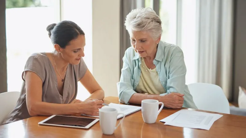 Questions to ask in-home care providers - Elderly woman sitting at table with female caregiver going over paperwork and drinking coffee